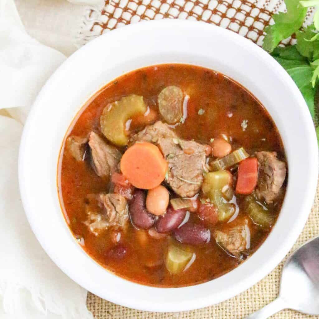 A bowl of beef and vegetable stew with carrots, celery, beans, and broth, placed on a table with a spoon and a white napkin nearby.