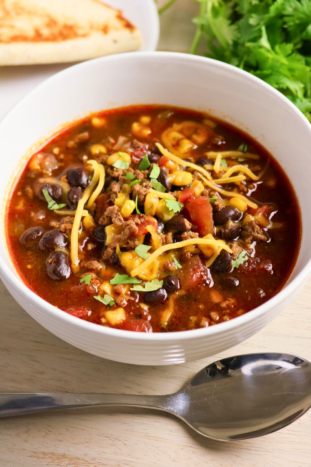 A bowl of chili with ground meat, black beans, corn, tomatoes, shredded cheese, and chopped herbs, placed next to a metal spoon on a light wooden surface.