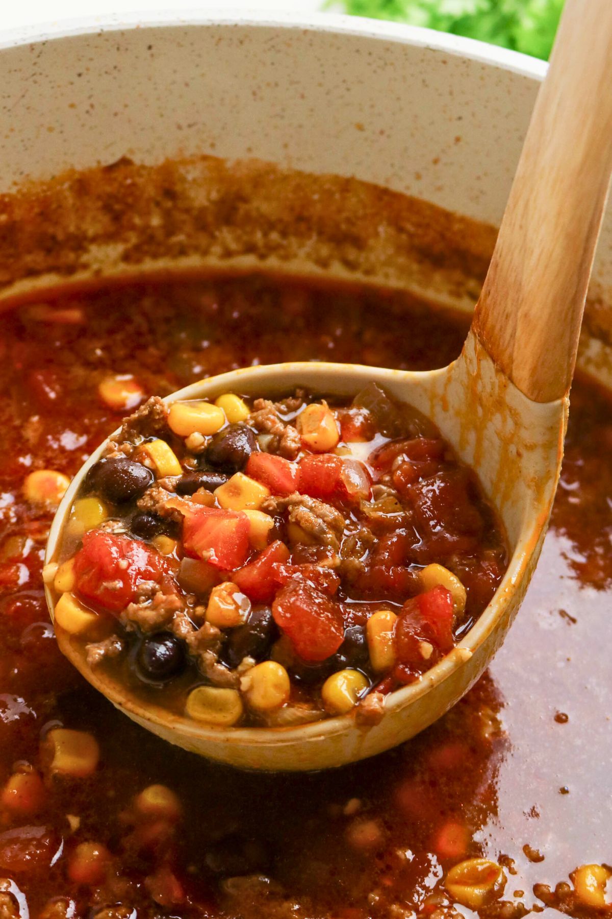 A close-up of a ladle lifting a serving of chili with ground meat, corn, black beans, and diced tomatoes from a pot.