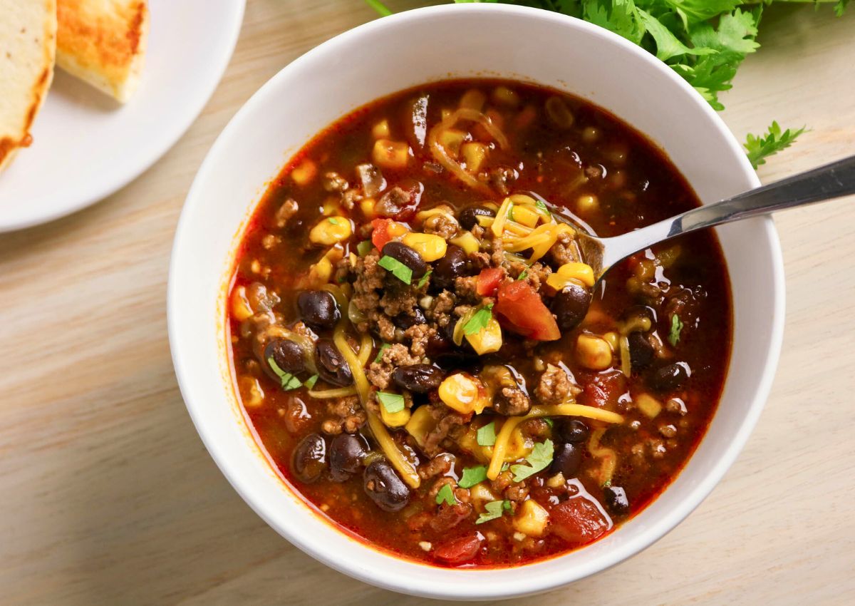 A bowl of chili with ground beef, black beans, corn, tomatoes, shredded cheese, and chopped herbs, with a spoon inside the bowl.