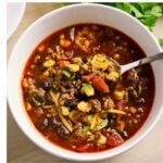 A bowl of chili with ground beef, black beans, corn, tomatoes, shredded cheese, and herbs, with a spoon inside the bowl.