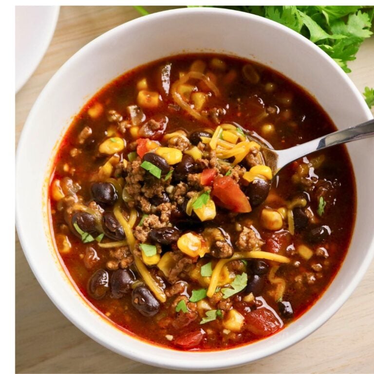 A bowl of chili with ground beef, black beans, corn, tomatoes, shredded cheese, and herbs, with a spoon inside the bowl.