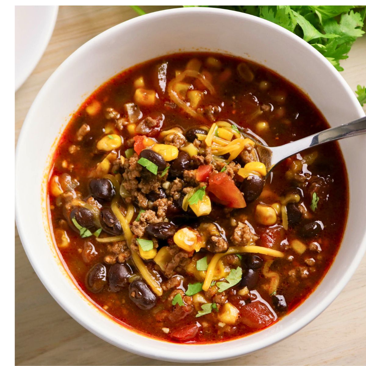 A bowl of chili with ground beef, black beans, corn, tomatoes, shredded cheese, and herbs, with a spoon inside the bowl.