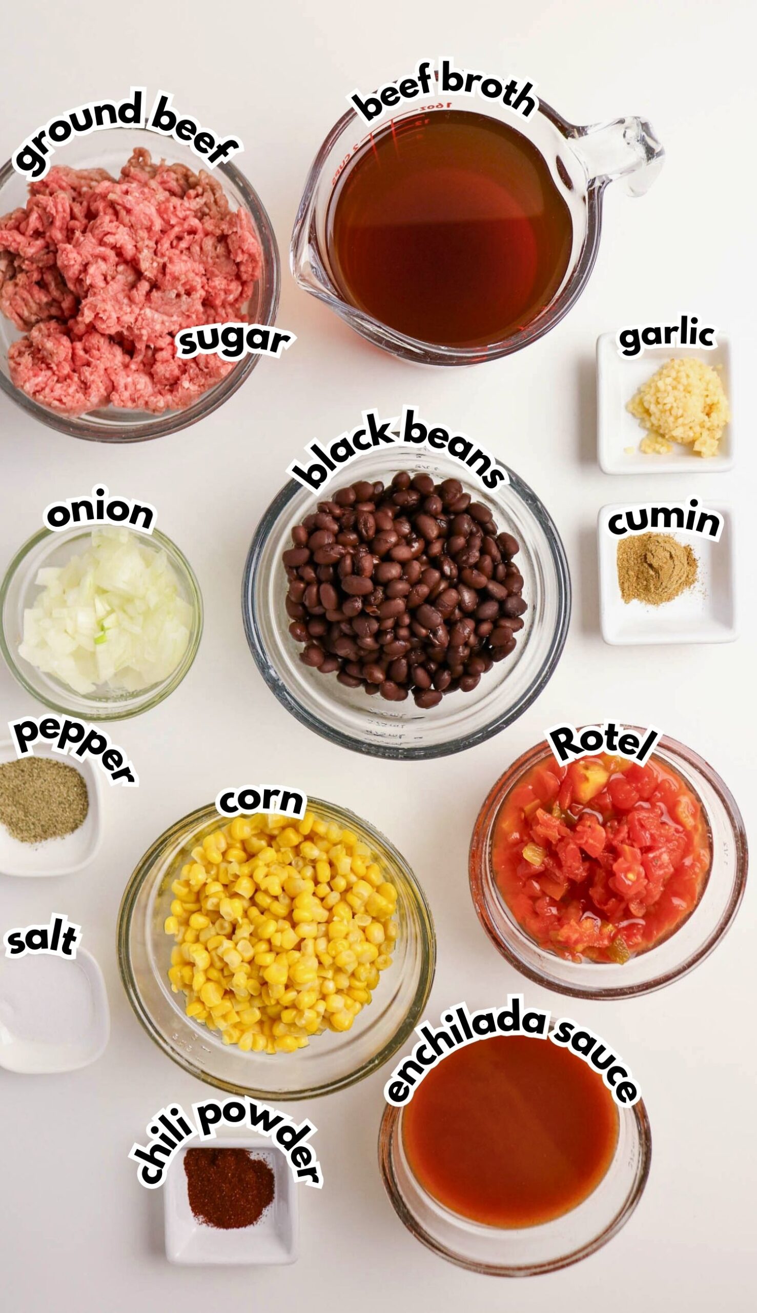 Ingredients for a recipe arranged on a white surface, including ground beef, black beans, corn, Rotel, enchilada sauce, beef broth, onion, garlic, and various seasonings.