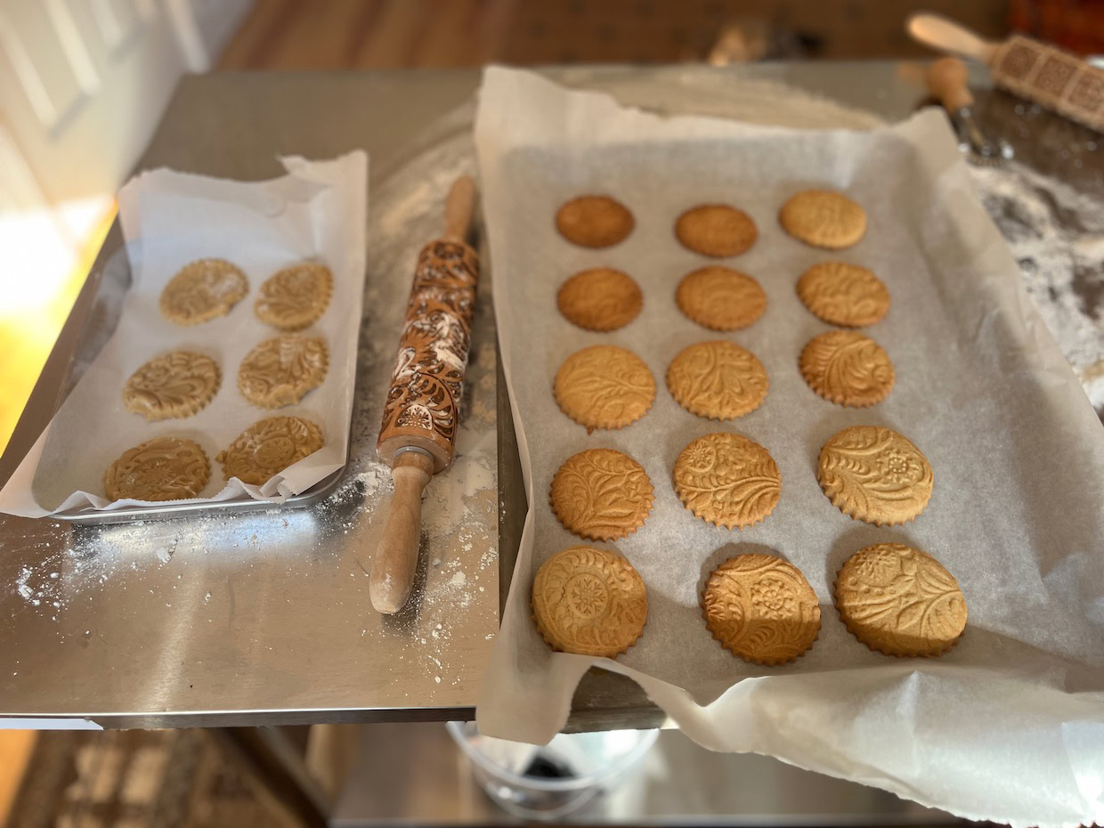 Two trays of embossed cookies on parchment paper sit on a metal surface; an engraved rolling pin with flour and dough is between them.