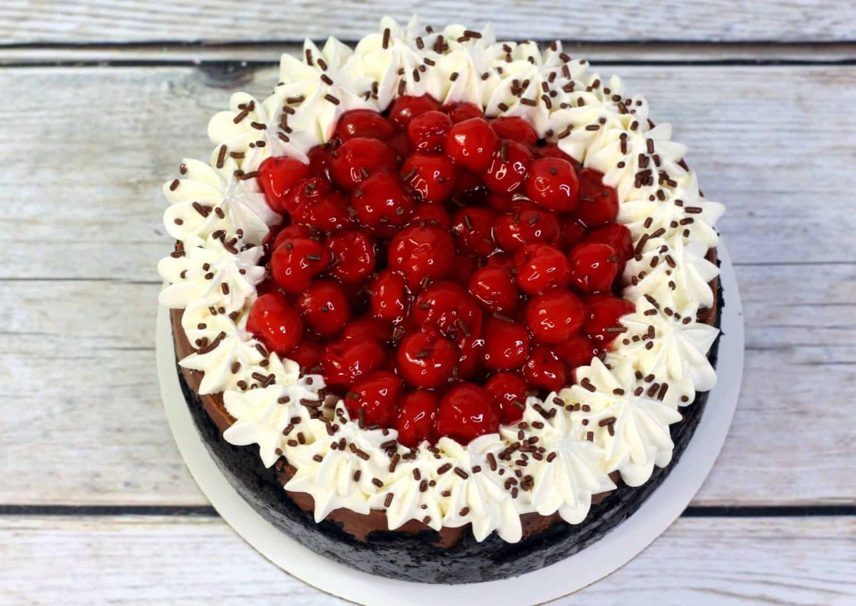 A chocolate cake topped with whipped cream, glazed cherries in the center, and chocolate sprinkles, placed on a wooden surface.