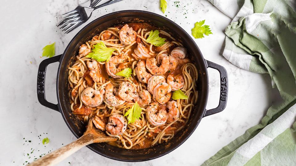 A pot of spaghetti with shrimp in tomato sauce, garnished with celery leaves, sits on a marble surface with a wooden spoon, forks, and a green-striped towel nearby.