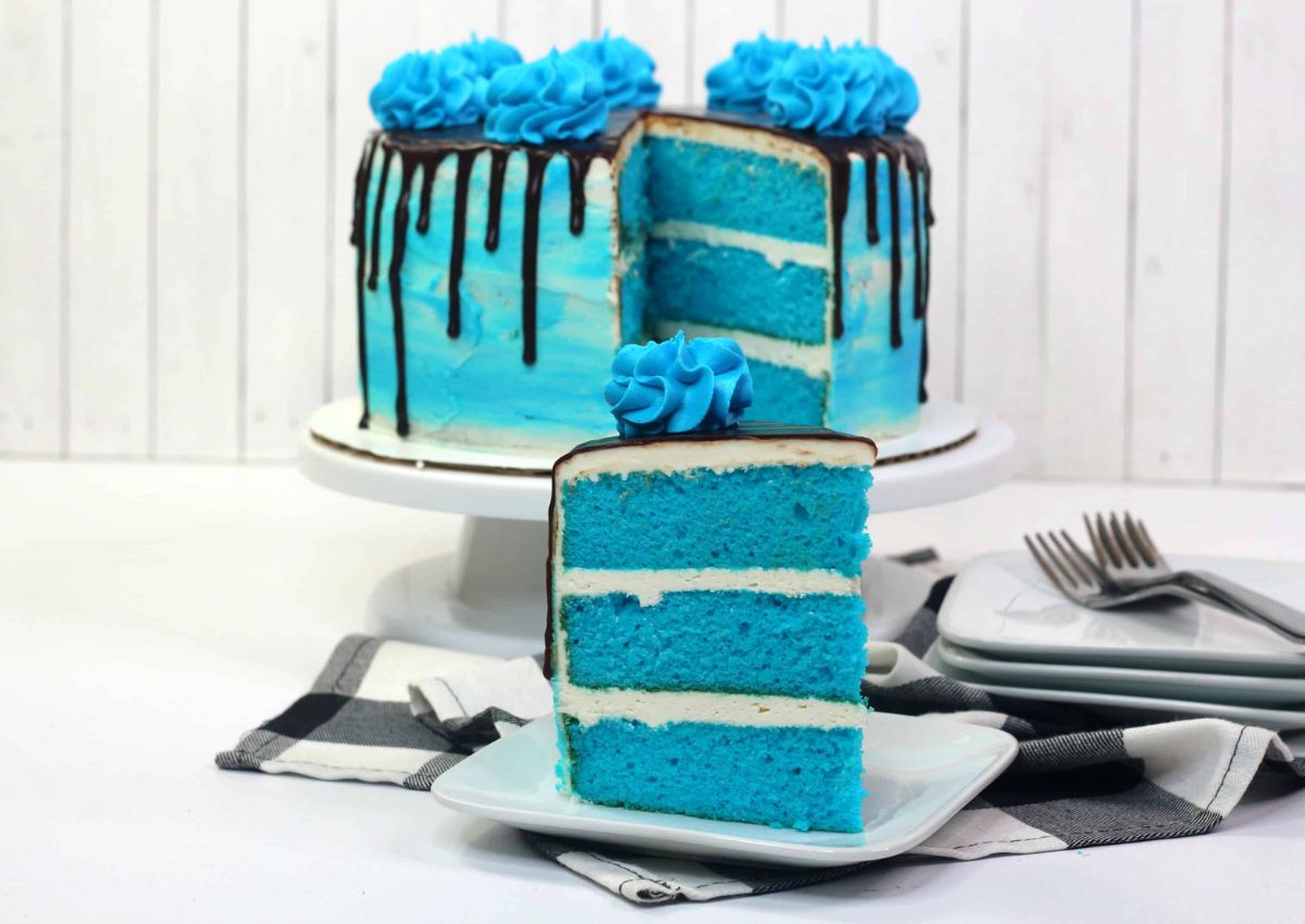 A three-layer blue cake with white frosting and blue icing sits on a stand. A slice is cut out and placed on a plate in the foreground. Three forks and stacked plates are nearby.