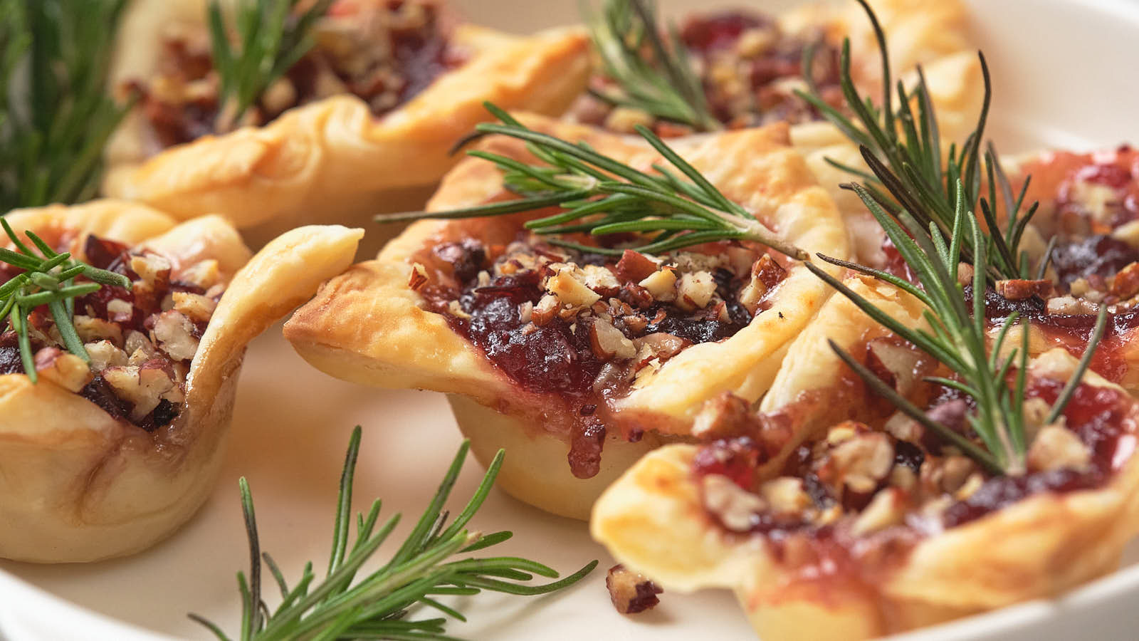 A close-up of pastry cups filled with a nut and cranberry mixture, garnished with fresh rosemary sprigs on a white plate.