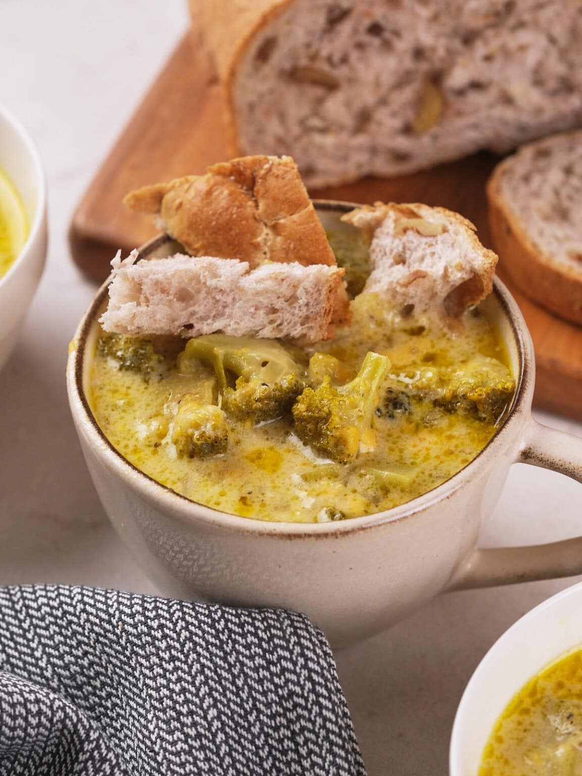A mug of broccoli soup topped with pieces of bread, with a sliced loaf of bread and a bowl of soup in the background. A patterned cloth is placed beside the mug.