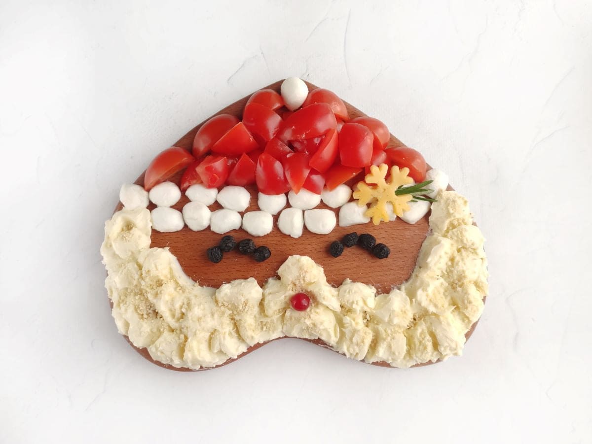 A heart-shaped wooden board decorated with chopped tomatoes, mozzarella balls, cheese, and other toppings to resemble Santa Clauss face.