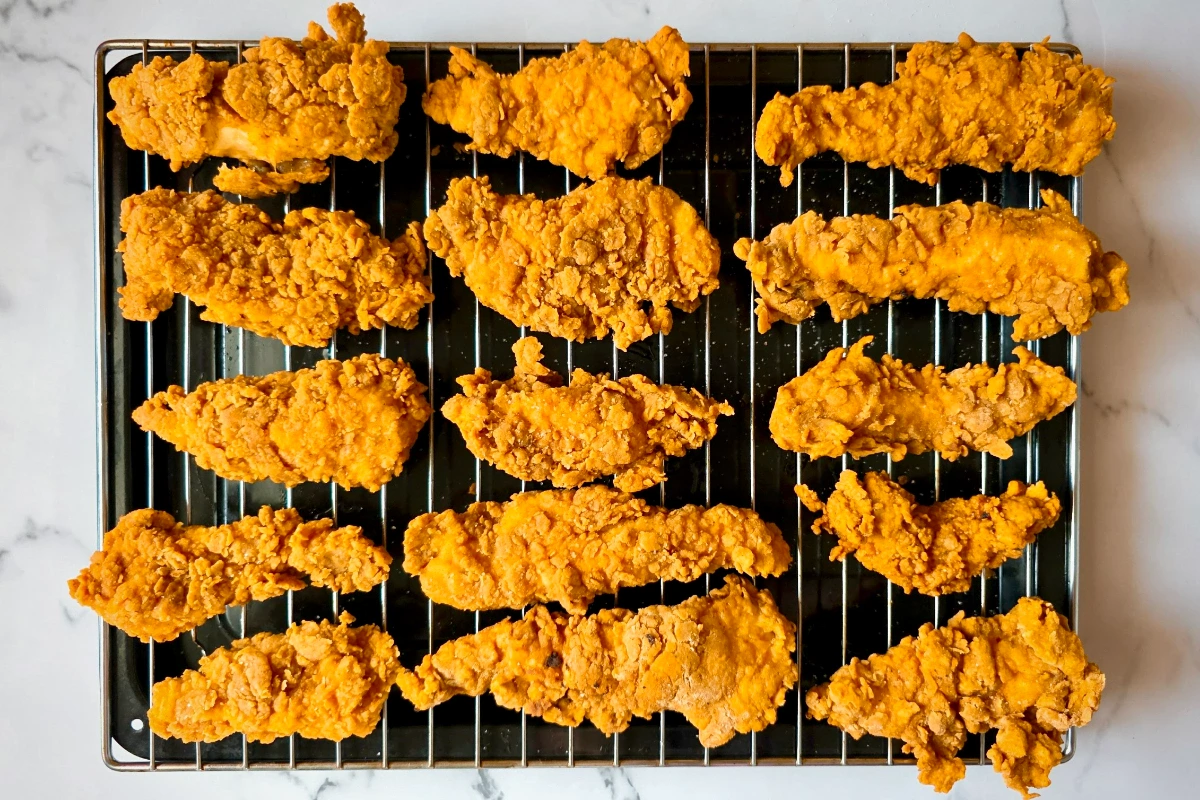 A wire rack on a baking sheet holds fourteen pieces of golden brown fried chicken strips arranged in rows.