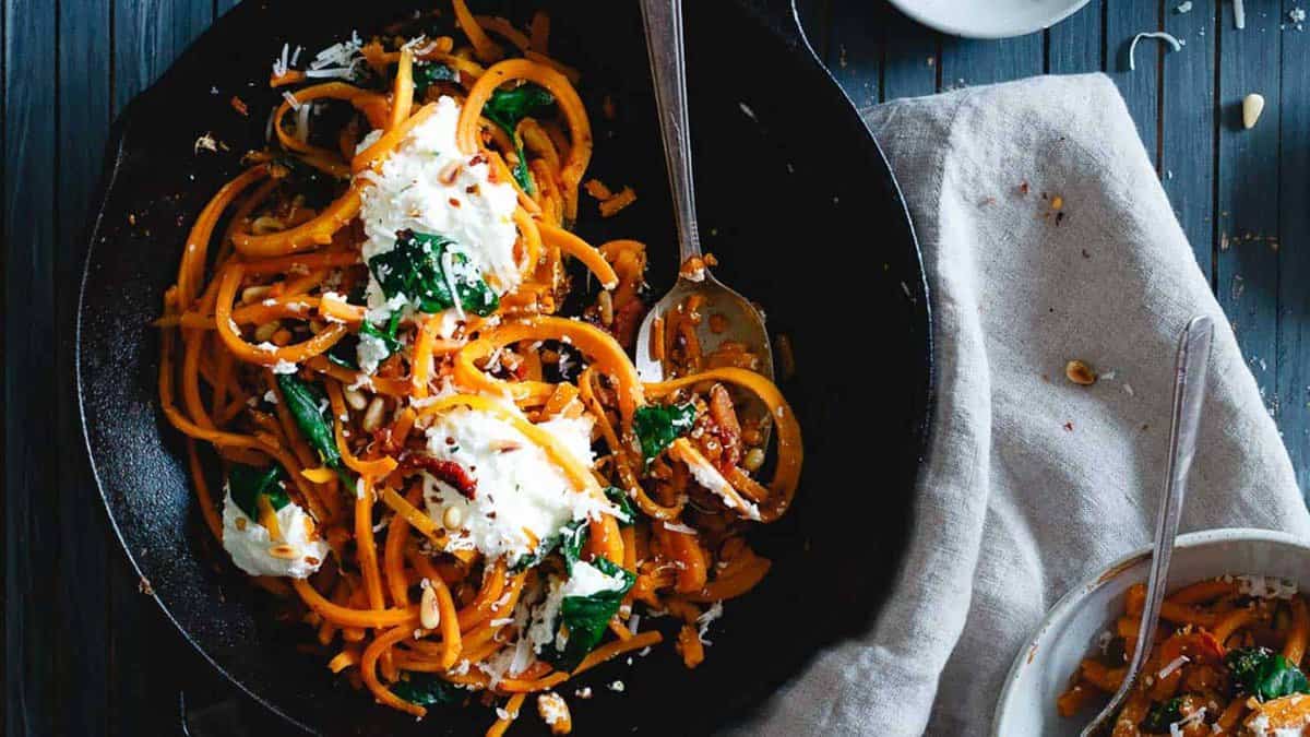 A skillet filled with spiralized sweet potatoes, wilted greens, crumbled cheese, and a fork, with a napkin and a small bowl on the side.