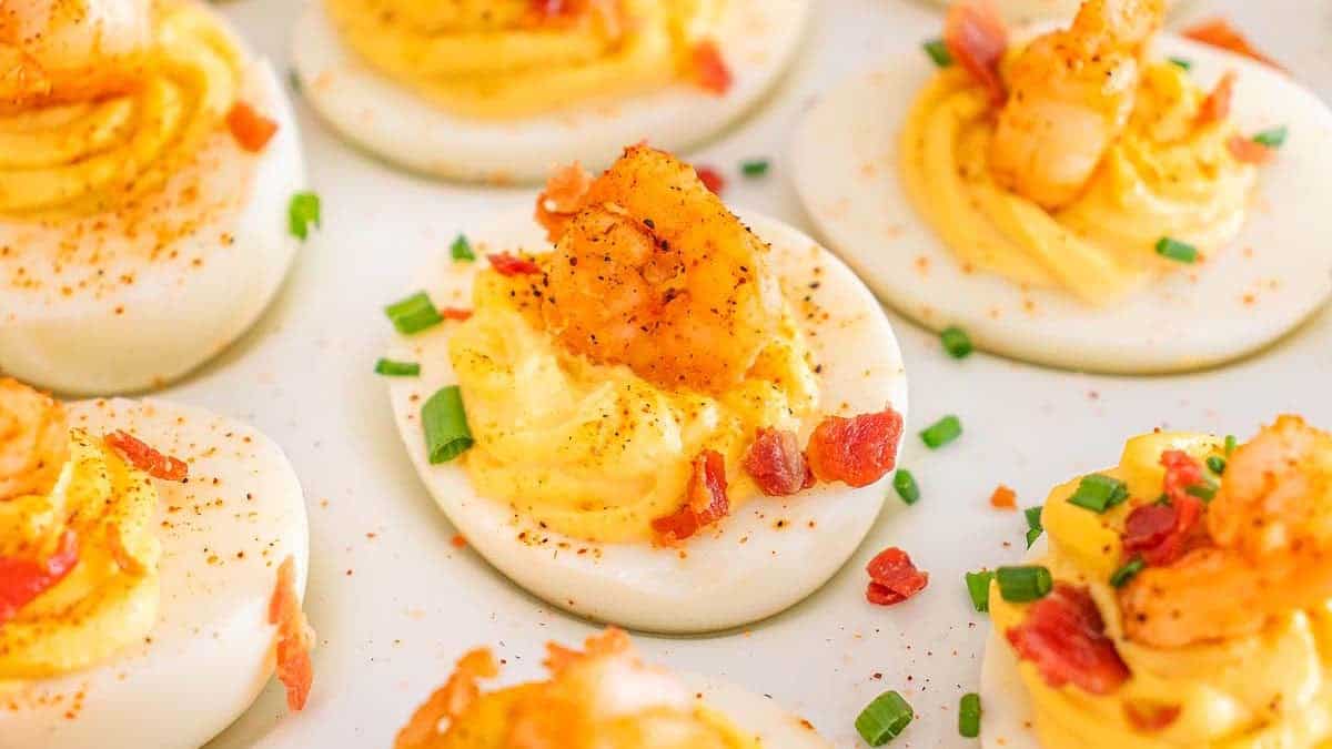 Deviled eggs topped with shrimp, chopped chives, bacon pieces, and a sprinkle of seasoning arranged on a white plate.