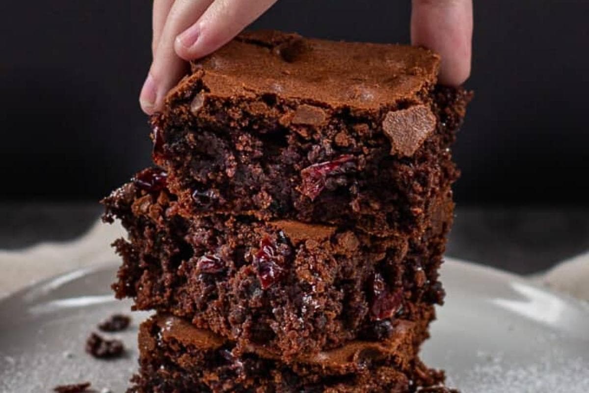 A hand holds the top piece of a stack of three chocolate brownies with visible chunks and dried fruit on a white plate.