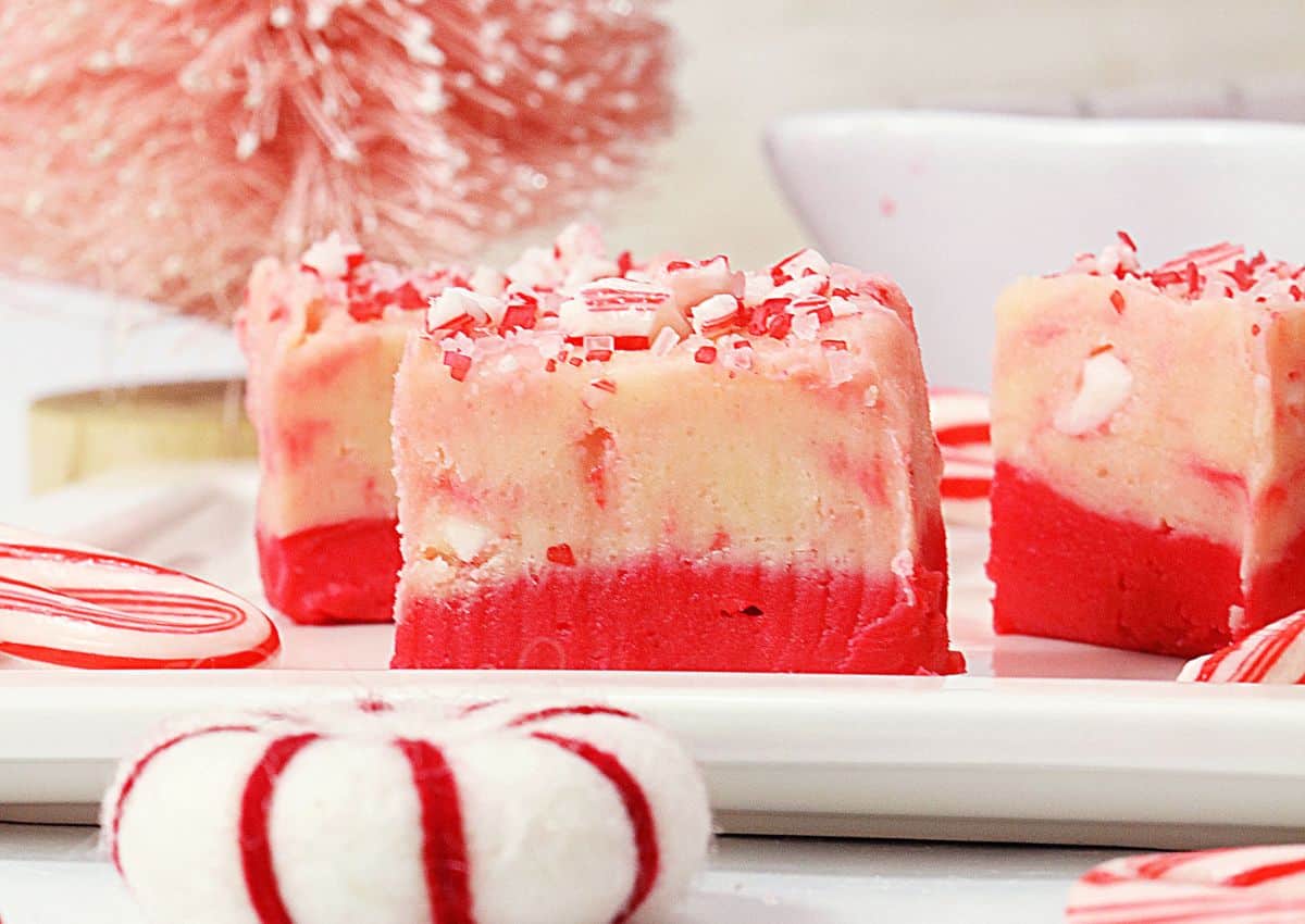 Three pieces of red and white layered peppermint fudge topped with crushed peppermint candy on a white plate, with festive decorations in the background.