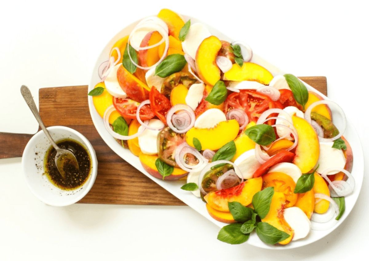 A platter of sliced tomatoes, peaches, mozzarella, onions, and basil leaves sits next to a small bowl of vinaigrette dressing on a wooden board.