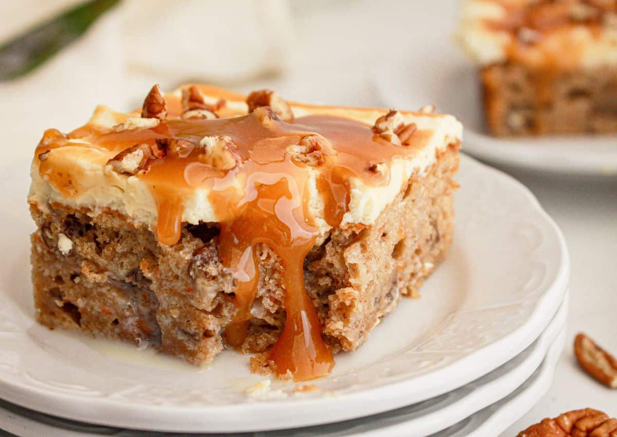 A slice of cake topped with creamy frosting, chopped pecans, and caramel sauce, served on a white plate.