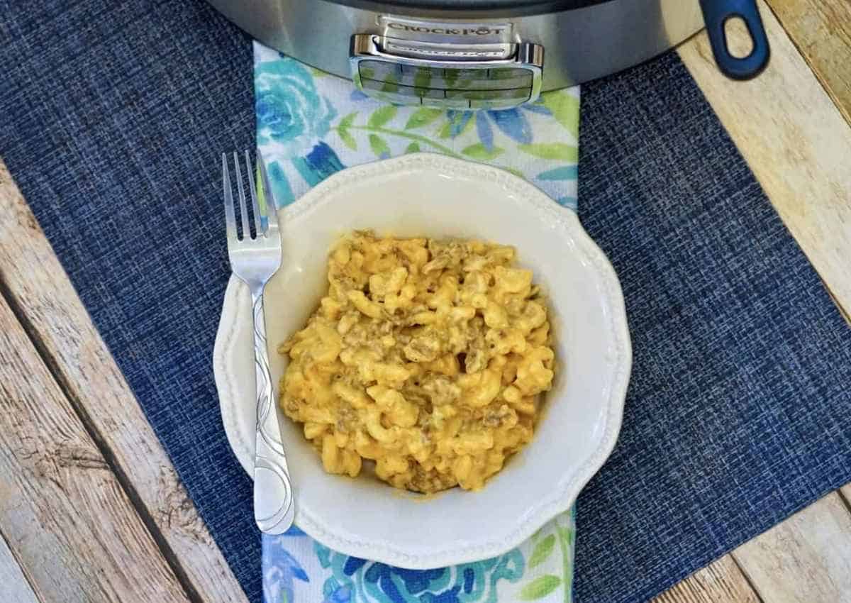 A white bowl of macaroni and cheese with ground meat sits next to a fork on a placemat, with a slow cooker visible in the background.