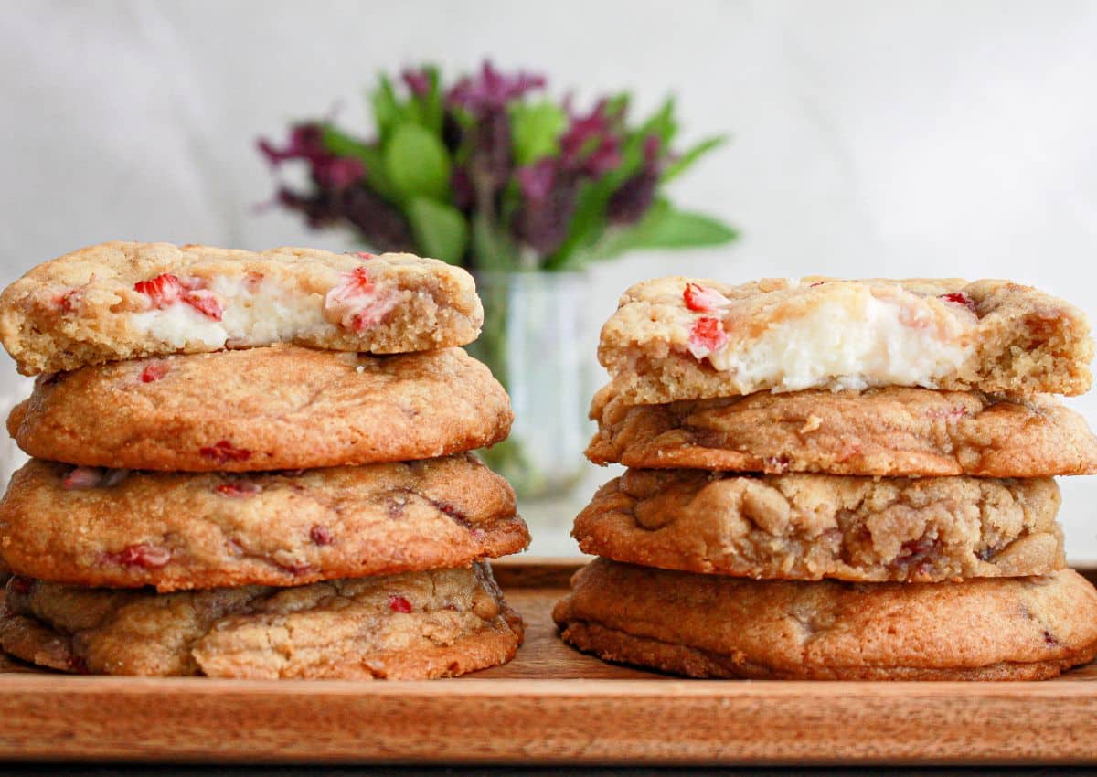 Two stacks of cookies with cream and red bits inside, displayed on a wooden board, with a blurred vase of flowers in the background.
