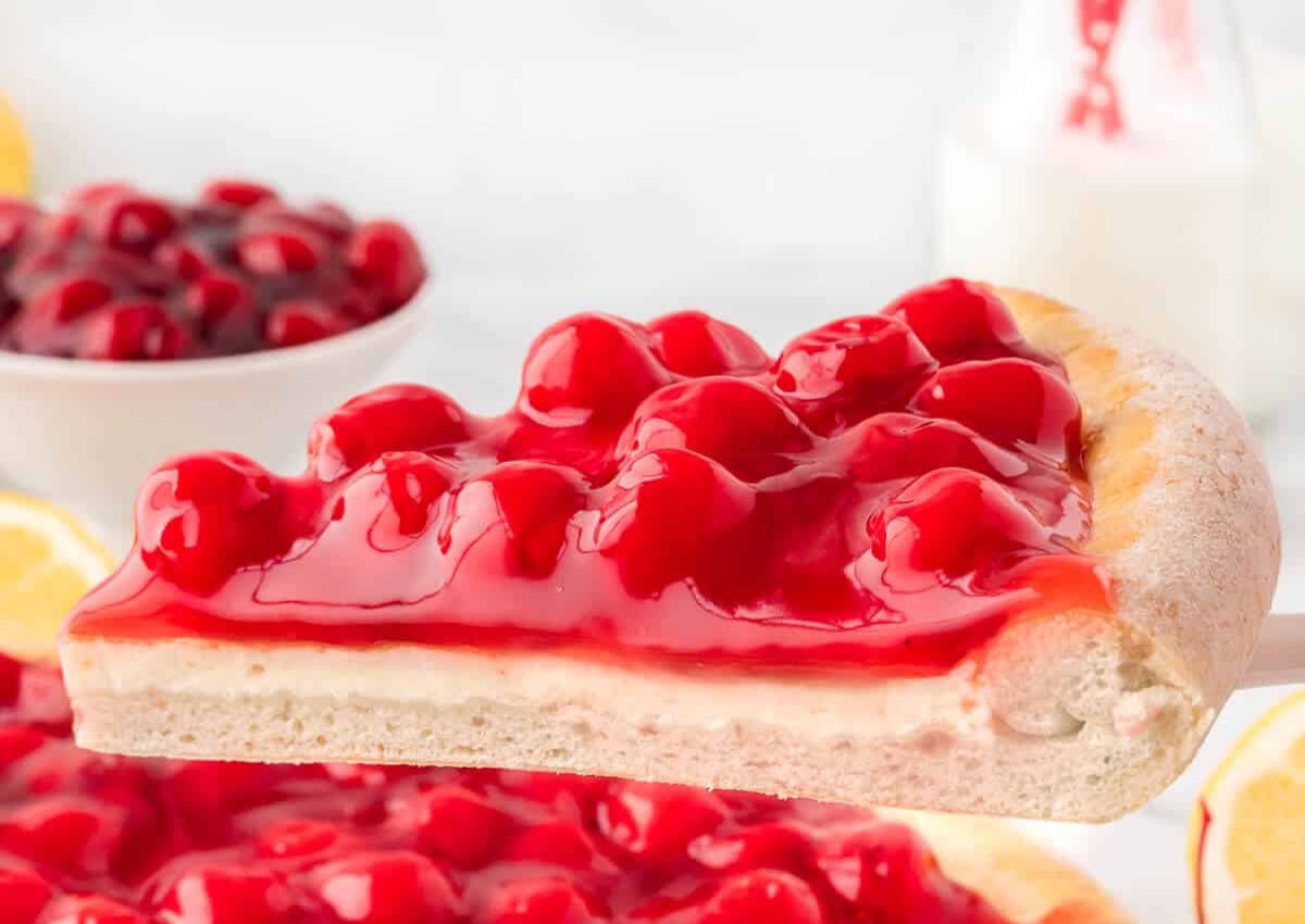A slice of cherry-topped dessert pizza with a thick crust and creamy base, held above a whole pizza, with cherries and milk in the background.