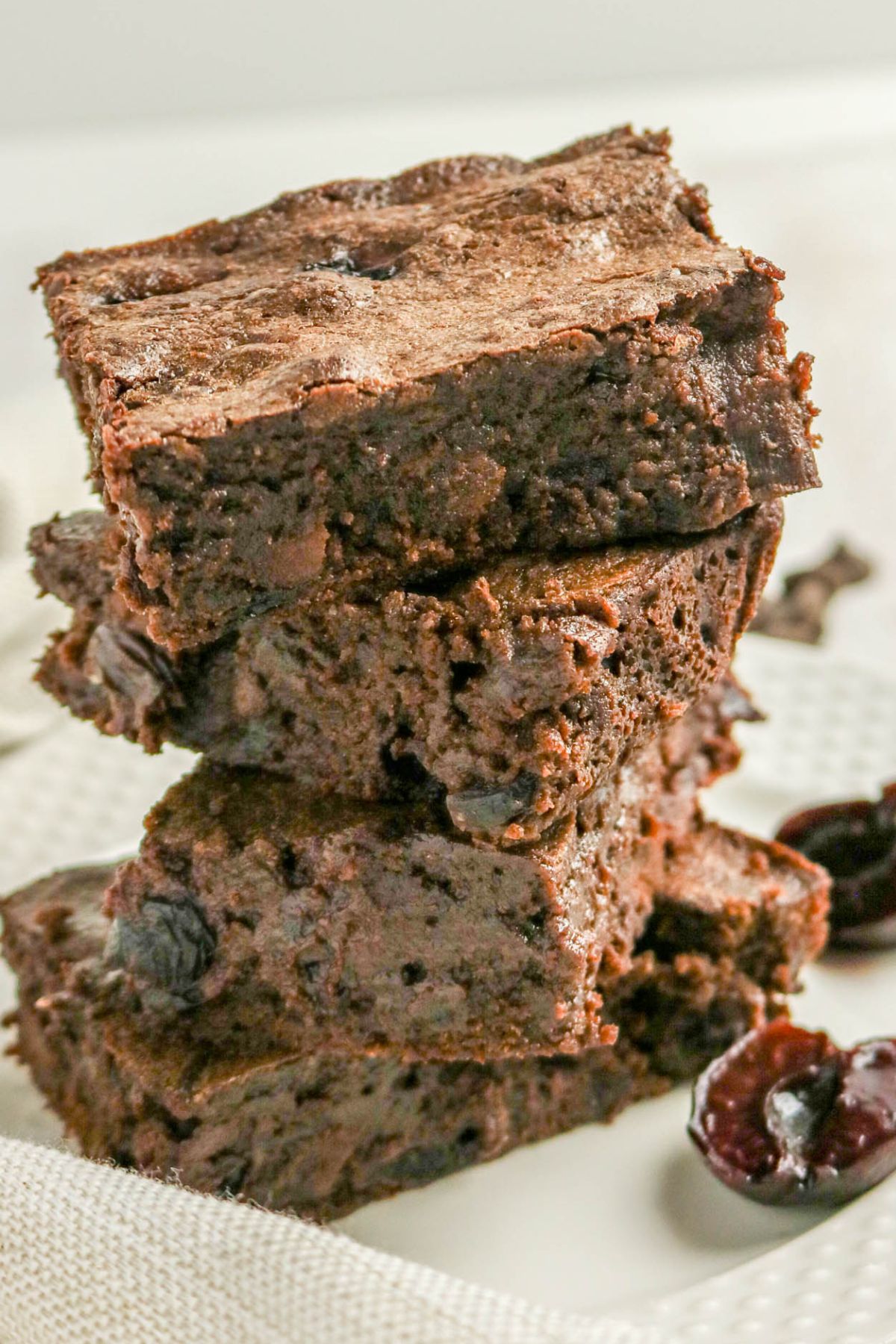 A stack of four thick, fudgy chocolate brownies with visible chocolate chunks, placed on a white textured surface.