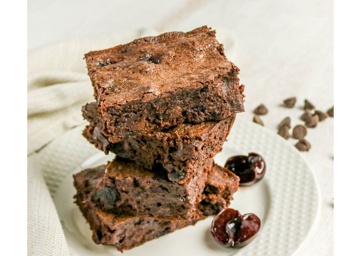 Three chocolate brownies stacked on a white plate next to halved cherries and scattered chocolate chips, with a beige cloth in the background.