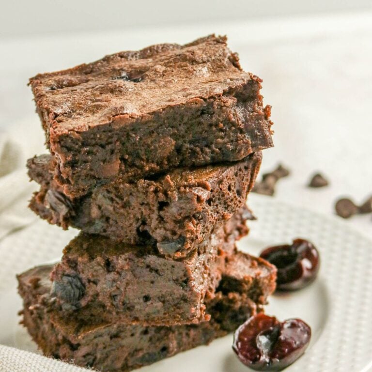 A stack of three chocolate brownies with visible chocolate chips, placed on a white plate next to halved cherries.