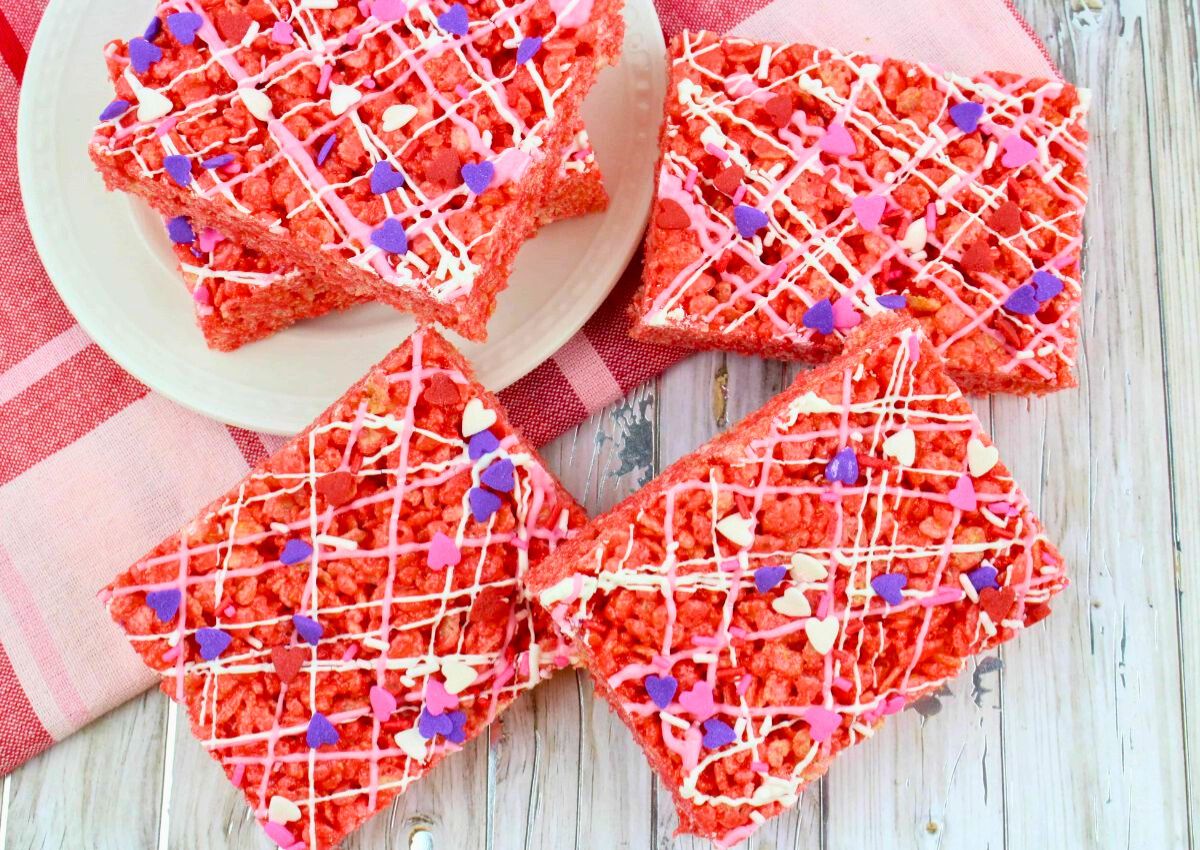 Four red rice krispie treats topped with white drizzle and pink, purple, and white heart-shaped sprinkles are arranged on a white plate and wooden surface.