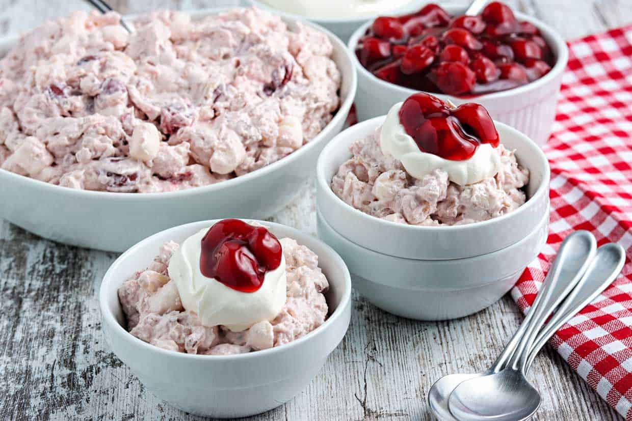 Three bowls of creamy fruit salad topped with whipped cream and cherries sit on a rustic table beside a larger serving bowl, with a red checkered cloth and spoons nearby.