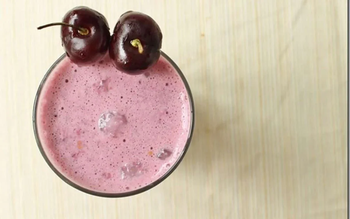 A glass of pink cherry smoothie with ice cubes, topped with two whole cherries, on a light wooden surface.