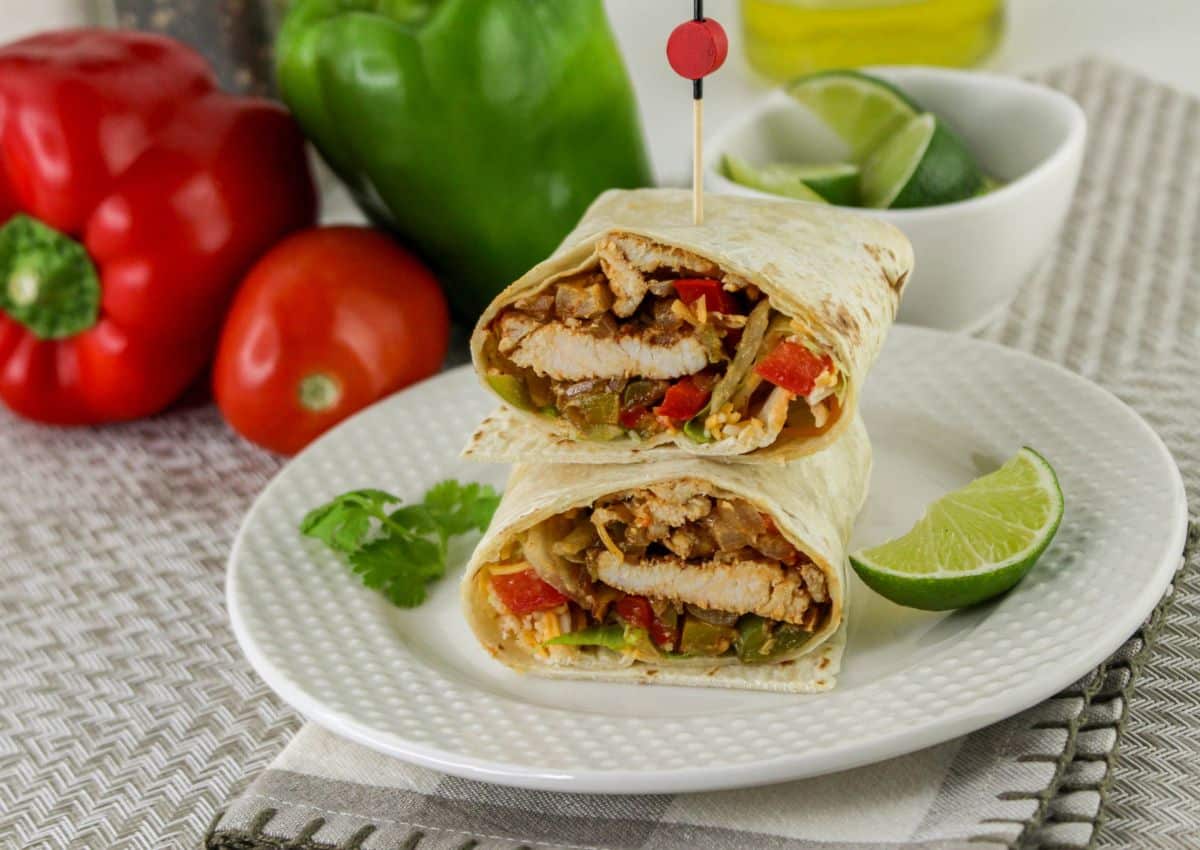 A plate with two halves of a chicken fajita wrap, garnished with lime and cilantro. A tomato, bell peppers, and a bowl of lime wedges are in the background.