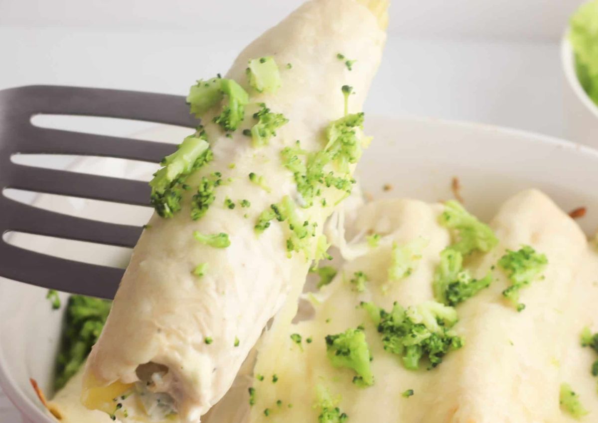 A spatula lifts a cheesy enchilada topped with chopped broccoli from a baking dish containing similar enchiladas.