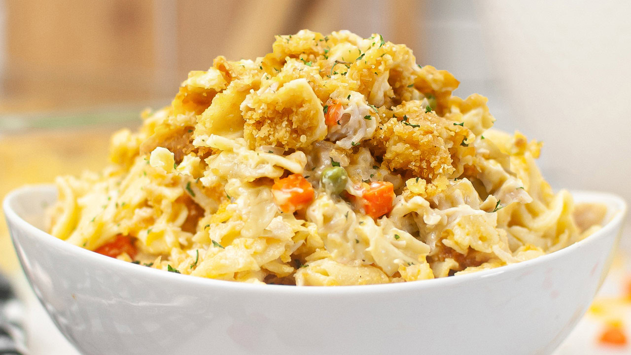 A white bowl filled with creamy pasta mixed with vegetables and topped with crispy breaded pieces and herbs.