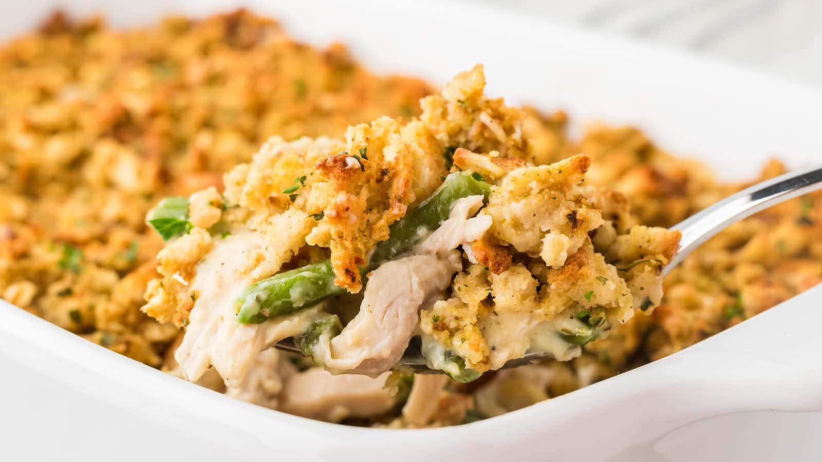 A close-up of a spoonful of casserole with green beans, shredded chicken, and a golden breadcrumb topping in a white baking dish.
