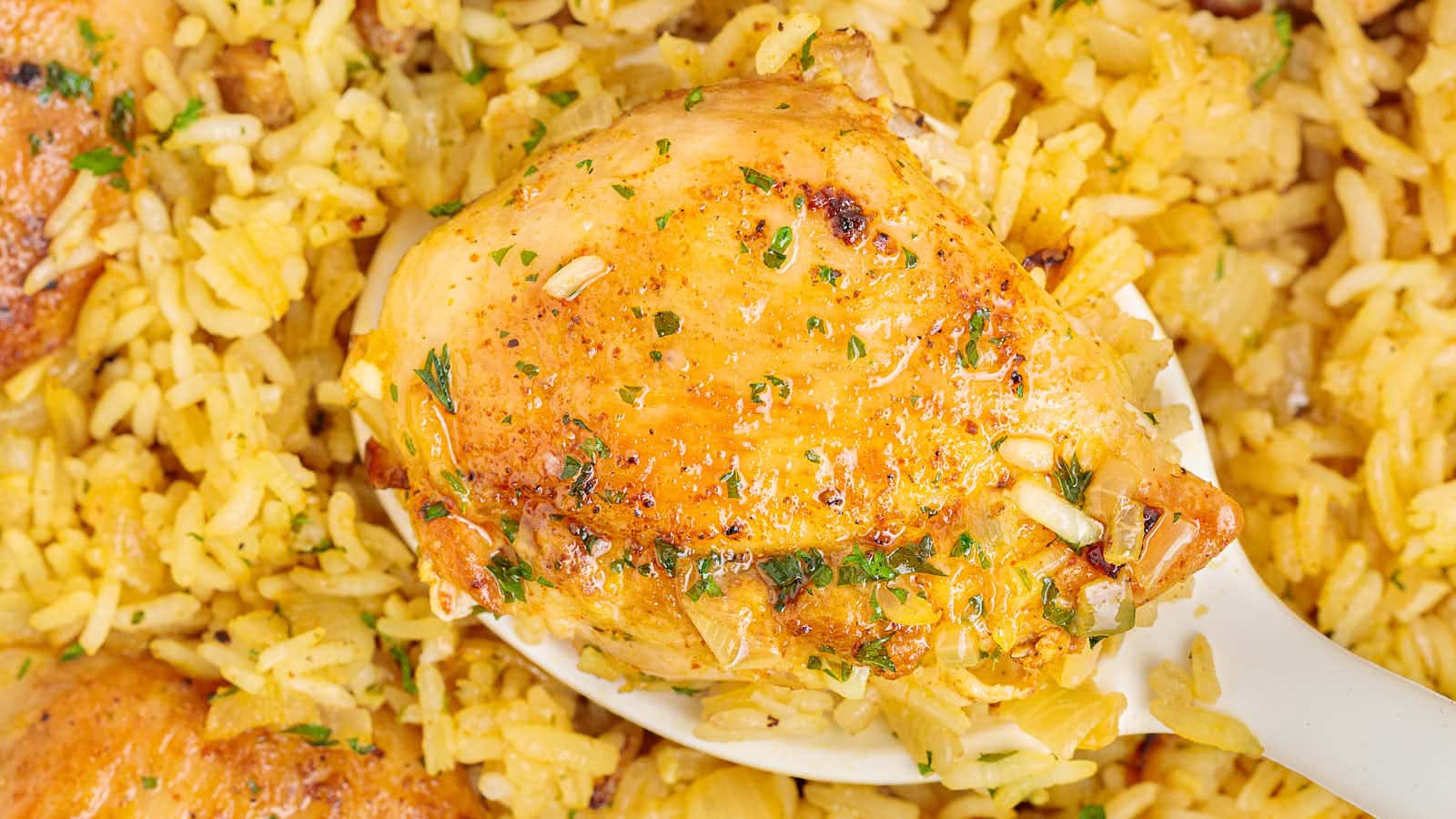 A close-up of a cooked chicken thigh with herbs on a spoon, served over a bed of seasoned yellow rice.