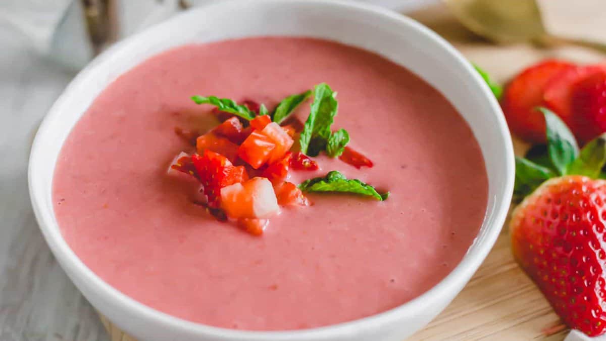 A white bowl filled with pink strawberry soup, garnished with chopped strawberries and mint leaves.