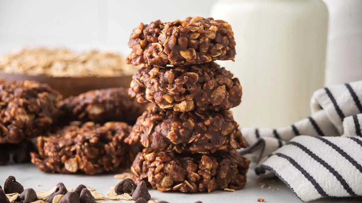 A stack of four no-bake chocolate oatmeal cookies on a white surface, with chocolate chips, oats, a striped towel, and a bottle of milk in the background.
