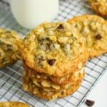 A stack of oatmeal chocolate chip cookies sits on a cooling rack with a glass of milk in the background.