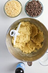 A mixing bowl with cookie dough and a paddle attachment, surrounded by bowls of white and milk chocolate chips, a bottle of vanilla extract, and a glass of water.