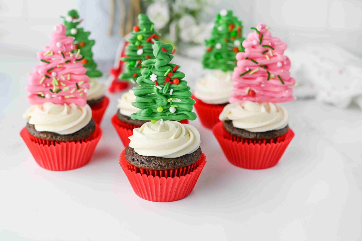 Chocolate cupcakes with white frosting in red wrappers, topped with pink and green Christmas tree decorations made of icing and sprinkles.