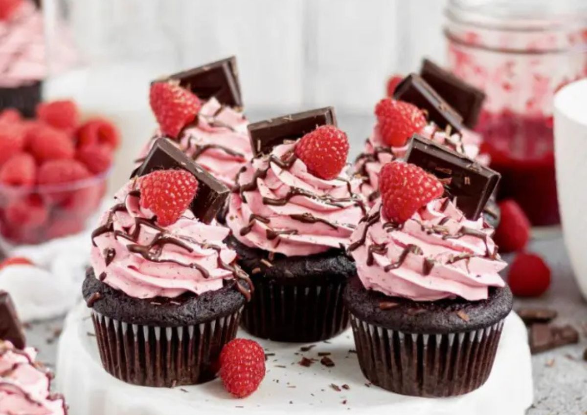 Chocolate cupcakes with pink raspberry frosting, topped with a raspberry, a piece of dark chocolate, and a drizzle of chocolate, displayed on a white plate.