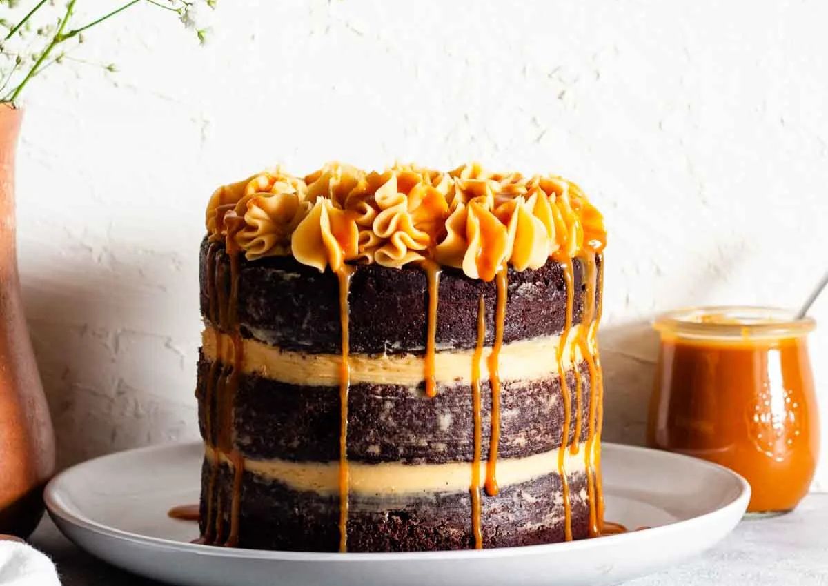 A tall, layered chocolate cake with caramel filling and frosting, topped with piped caramel cream and caramel sauce dripping down the sides, sits on a white plate.
