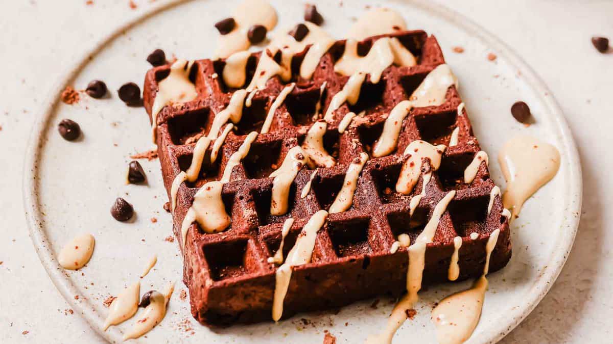 A chocolate waffle drizzled with sauce sits on a white plate, with chocolate chips and sauce splatters around it.