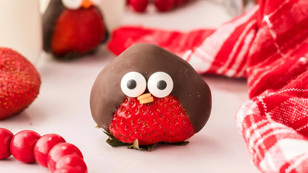 A strawberry coated with chocolate, decorated with candy eyes and a beak to resemble a bird, sits on a white surface near a red checkered cloth.