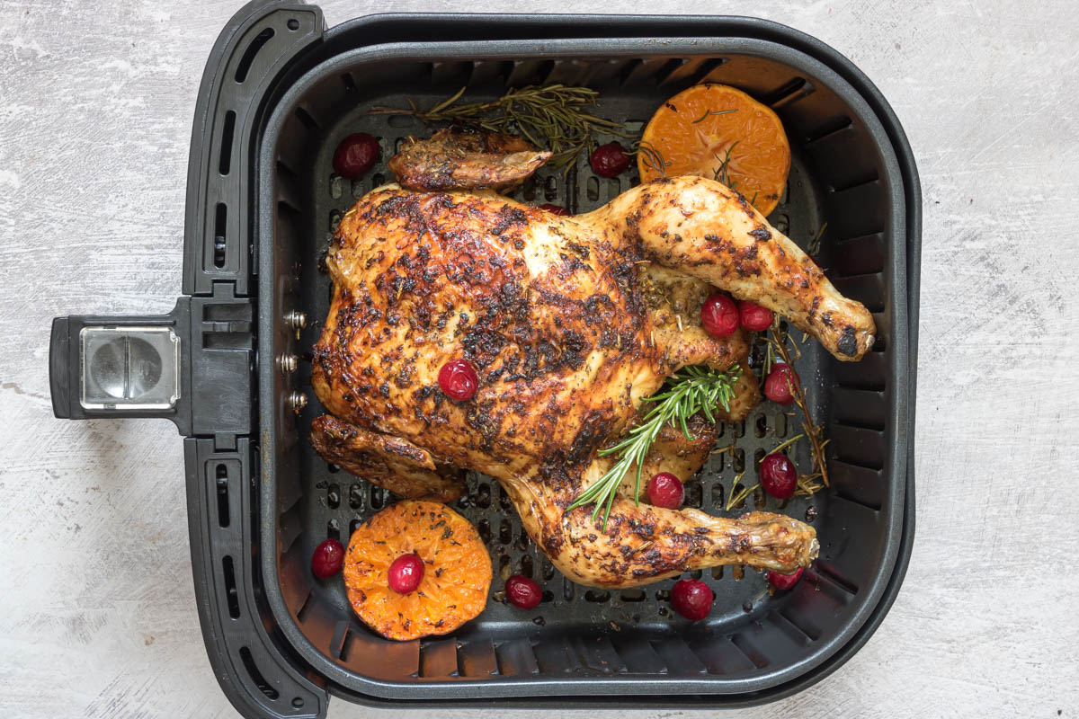 A whole roasted chicken with herbs, orange slices, and cranberries in an air fryer basket.