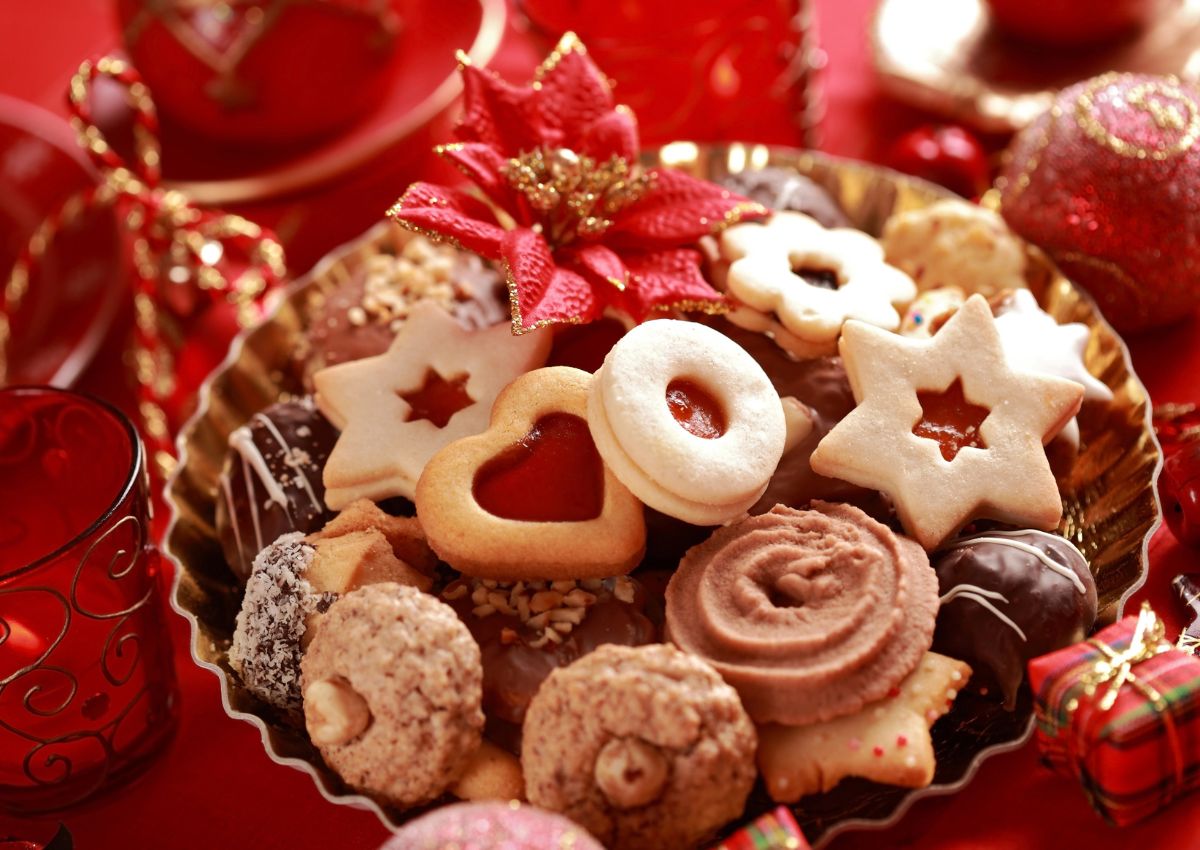A gold plate filled with assorted Christmas cookies, including jam-filled, star-shaped, and chocolate-dipped varieties, on a festive red background with ornaments.