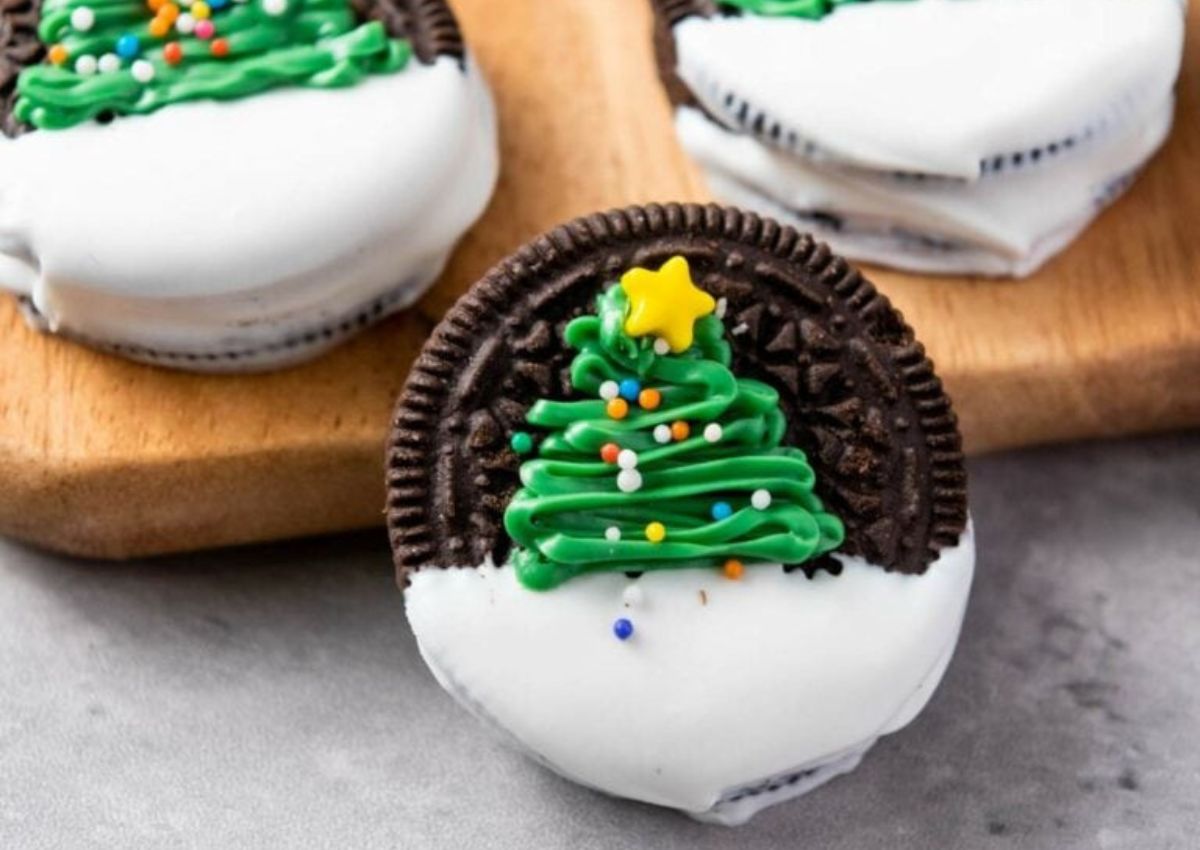 A chocolate sandwich cookie half-dipped in white coating, decorated with green icing shaped like a Christmas tree and multicolored sprinkles.