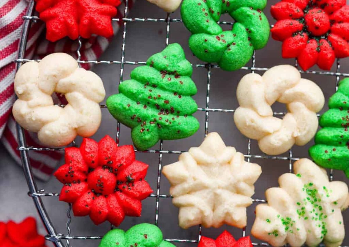 Christmas-themed spritz cookies in red, green, and white, shaped like wreaths, trees, poinsettias, and snowflakes, cooling on a wire rack.