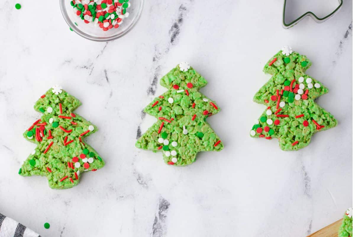 Three green Christmas tree-shaped cookies with red, white, and green sprinkles are arranged on a white marble surface.